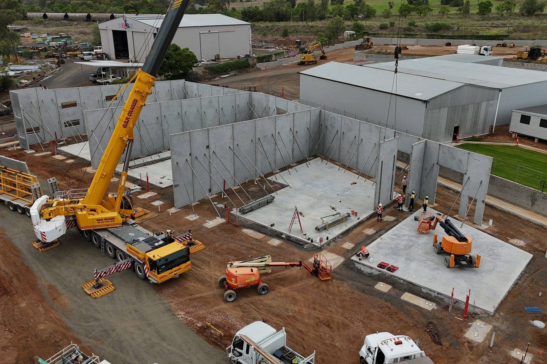 Aerial view of an active commercial development site under construction — tilt-up concrete panels being erected by crane, with cherry pickers, ground crew, and supply trucks in frame